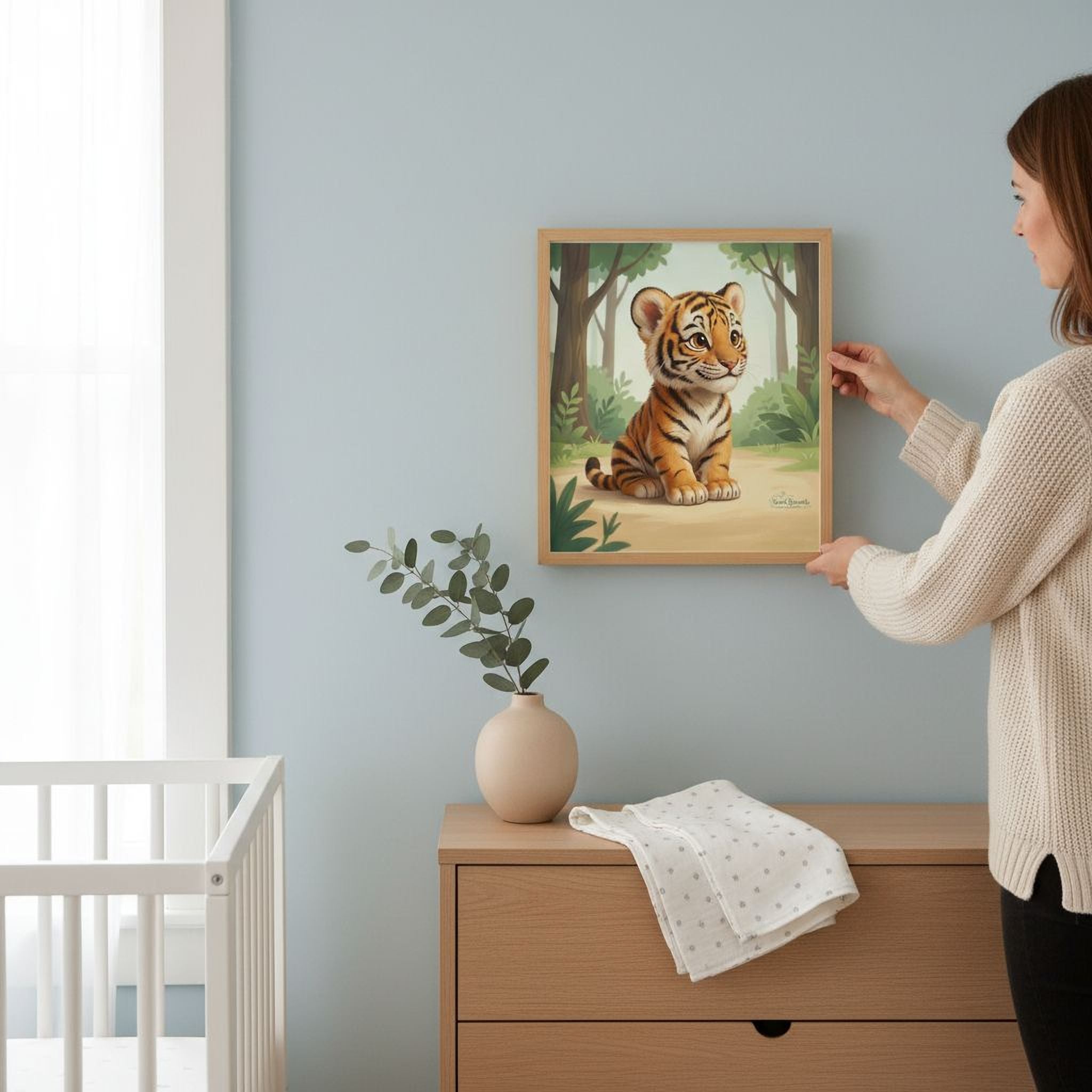 A woman is hanging a framed painting of a tiger on a wall in a room with a crib, a vase with greenery, and a wooden dresser.
